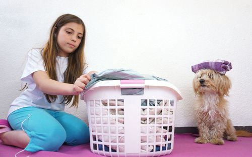 Girl folding clothes in laundry basket and dog balancing towel against white wall