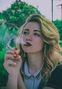 Young woman smoking cigarette while looking away against sky