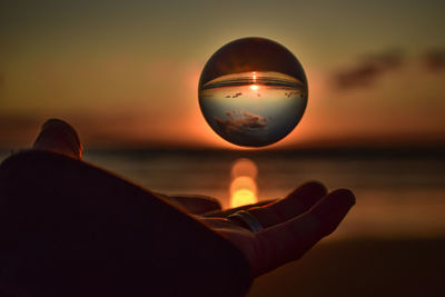 Man hand holding crystal ball against sea during sunset