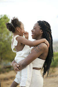 Side view of glad african american mom in white clothes with braided hair smiling and embracing daughter while standing against lush trees and cloudy sky in countryside