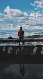 Rear view of man standing at lakeshore against cloudy sky