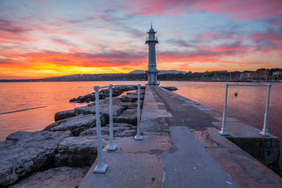 Walkway amidst lake leading towards lighthouse against sky during sunset