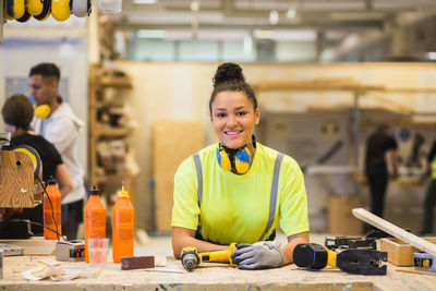 Smiling young female trainee leaning on workbench at illuminated workshop