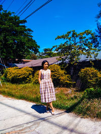 Full length of young woman looking away while standing on footpath against blue sky