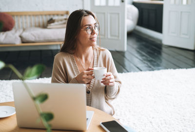 Young smiling woman in beige cardigan and jeans using laptop drinking coffee in room