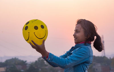 Girl holding balloon with smily printed against sky during sunset