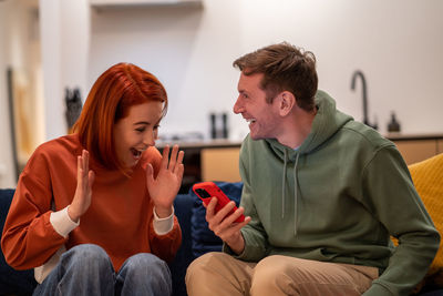 Happy overjoyed couple man woman reading good news in smartphone sitting on couch at home.