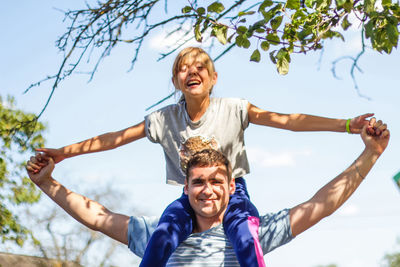 Portrait of smiling young man with arms raised standing against trees