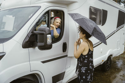 Senior woman sitting camper van talking to young woman standing with umbrella in rain