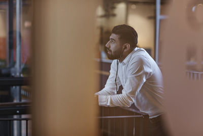 Young businessman listening through headphones while standing at railing in office