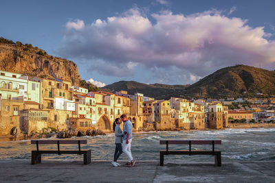 Woman standing on bench by buildings against sky