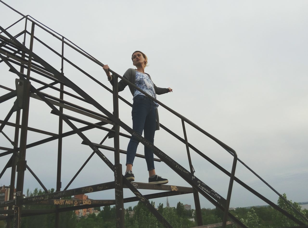 lifestyles, leisure activity, low angle view, young adult, casual clothing, standing, full length, clear sky, young men, sky, copy space, person, built structure, connection, men, day, three quarter length, outdoors