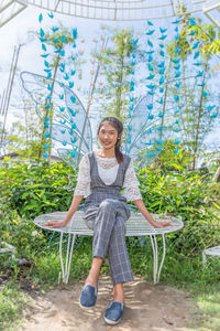 Full length portrait of smiling young woman sitting on butterfly structure against sky