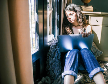 Young woman using phone while sitting on window