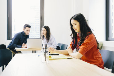 Businesswoman examining documents while coworkers discussing over laptop computer in background at office