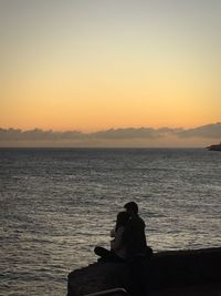 Rear view of silhouette man sitting on shore against sunset sky