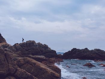 Person walking on rock formation at beach against cloudy sky