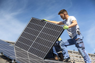 Low angle view of worker installing solar panel on roof against sky