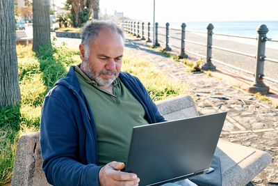 Handsome bearded mature man with laptop working outdoors while sitting on bench at the seaside. 