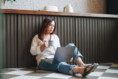 Adult brunette middle aged woman in casual clothes with morning coffee using laptop at home