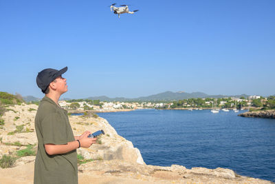 Happy teen boy, flying drone on mediterranean coast, against blue sky during sunny day mallorca