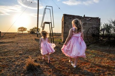 Full length rear view of sisters walking on field against sky during sunset