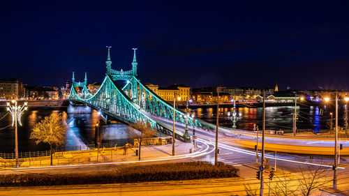 Light trails on bridge over river against sky in city at night