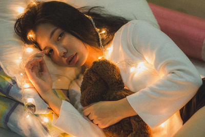 Portrait of young woman with string lights and toy lying in bed