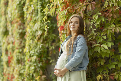 White pretty pregnant woman stand near green overgrown liana wall,looks at camera,healthy female