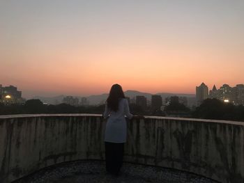 Rear view of woman standing by railing against sky during sunset