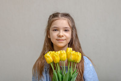 A girl in a blue dress with yellow tulips in her hands against a background of a gray wall