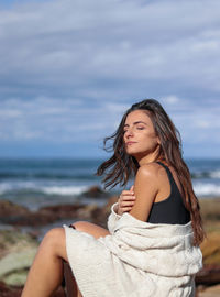 Portrait of young woman sitting at beach against sky