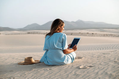 Back view of content young female tourist with tablet sitting on sandy dunes of corralejo against straw hat and mount on sunny day in fuerteventura canary islands spain