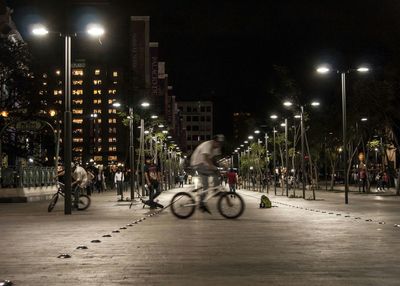 Boys riding bicycle on road amidst illuminated street lights during night