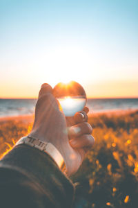 Close-up of hand holding crystal ball against sky during sunset