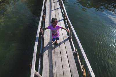 High angle view of girl with arms outstretched walking on footbridge over lake
