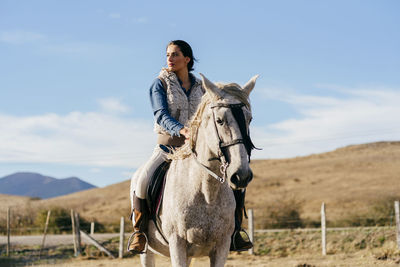 Woman in hat riding a beautiful white horse on background of rural landscape under blue sky