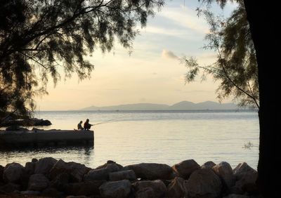 Silhouette people sitting by sea against sky during sunset