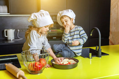 Siblings wearing chef's hat eating apple fruit in kitchen
