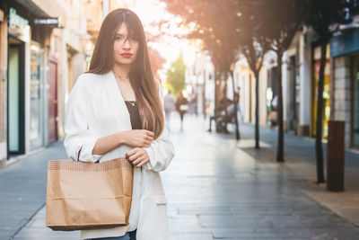 Portrait of young woman with shopping bag standing on footpath in city