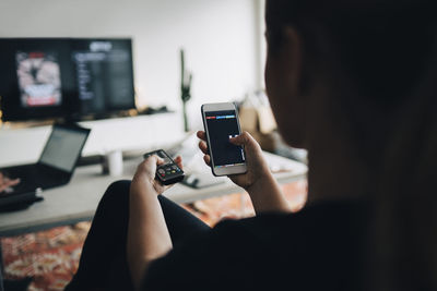 Rear view of teenage girl using phone app and remote control while watching smart tv at home