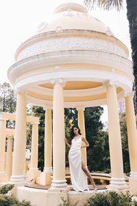 A beautiful brunette lady in an elegant wedding dress poses among the columns in the old city park