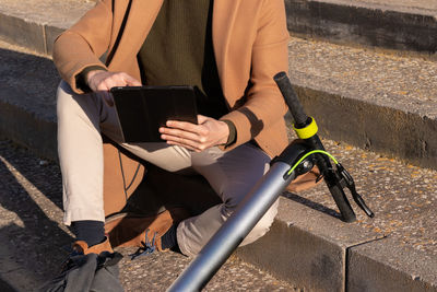 Cropped close up view of unrecognizable man sitting in the park with tablet and electric scooter 