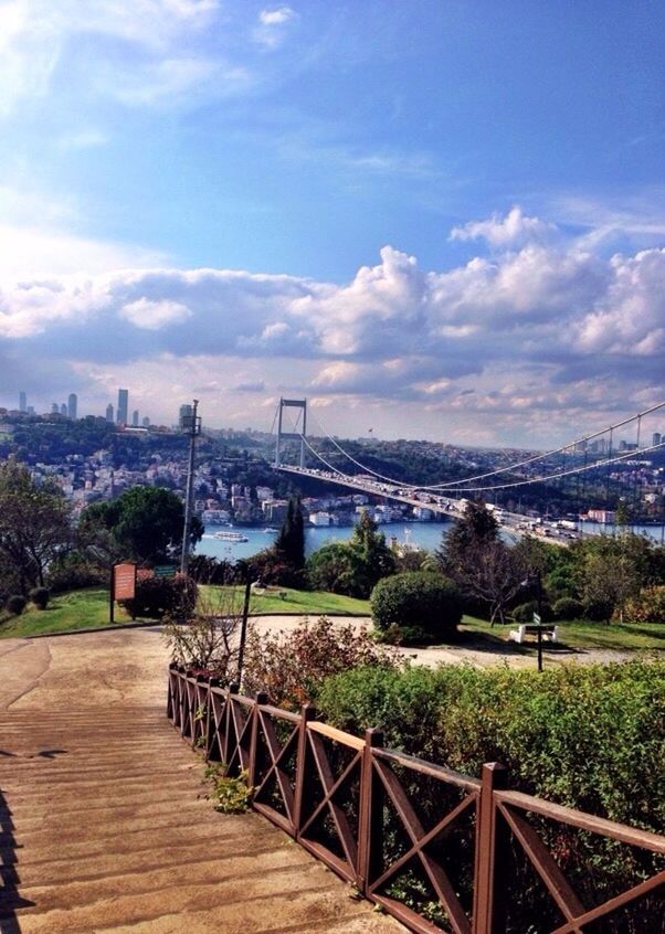 built structure, architecture, sky, building exterior, railing, tree, city, cloud - sky, bridge - man made structure, cloud, cityscape, connection, fence, river, walkway, day, high angle view, the way forward, grass, outdoors