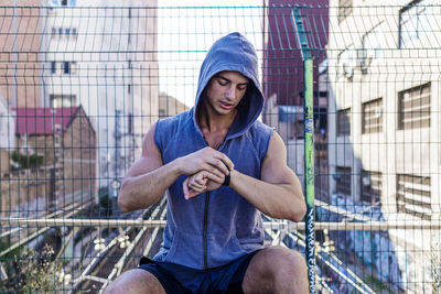 Muscular man checking time while sitting against fence