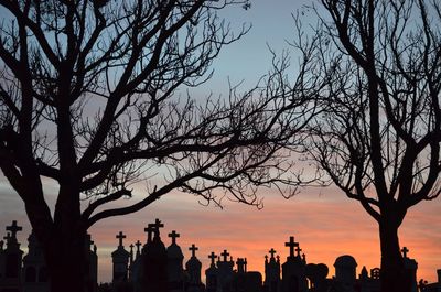 Silhouette crosses on tombstones by bare trees at cemetery against sky during sunset