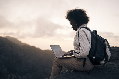 Side view of female backpacker using laptop while sitting on rock at sunset