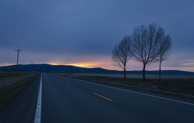 Road by bare trees against sky during sunset
