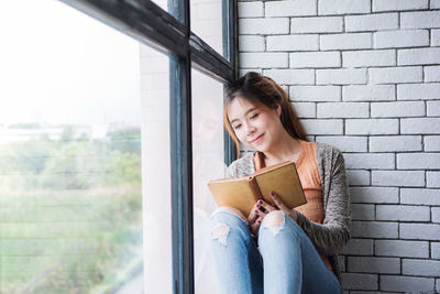 Young woman reading book while sitting on window