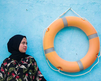 Young woman wearing hijab looking at life belt hanging on blue wall